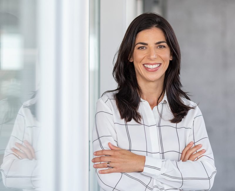 Portrait of middle aged businesswoman in modern office looking at camera. Confident business woman with arms crossed standing while leaning against glass wall. Proud brunette woman smiling in formalwear with copy space.