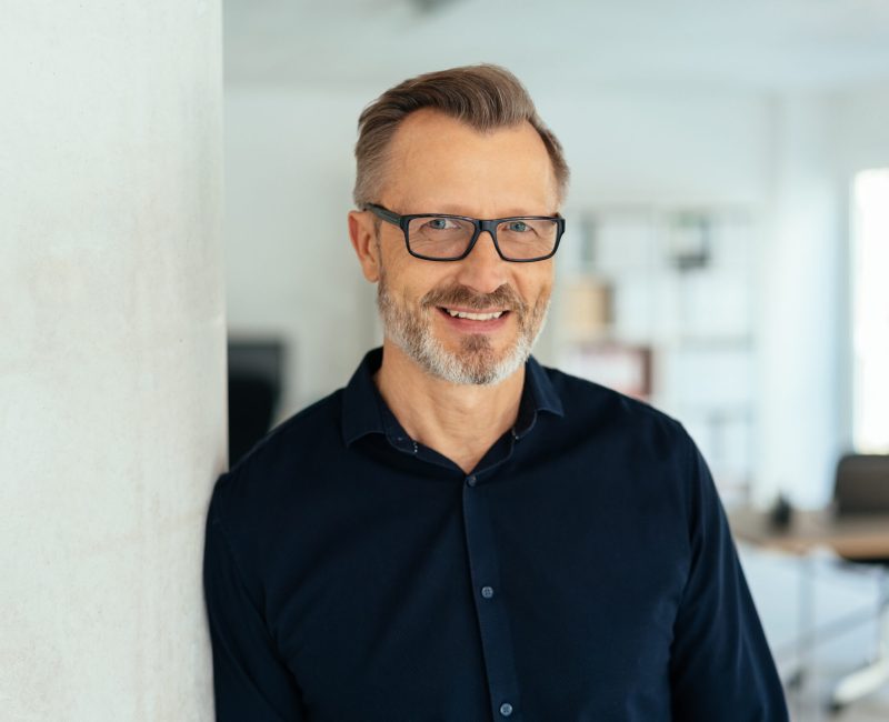 Indoor close-up portrait of a smiling handsome bearded middle-aged man in glasses and black shirt, leaning with his shoulder on white wall and looking at camera with friendly face, standing in office