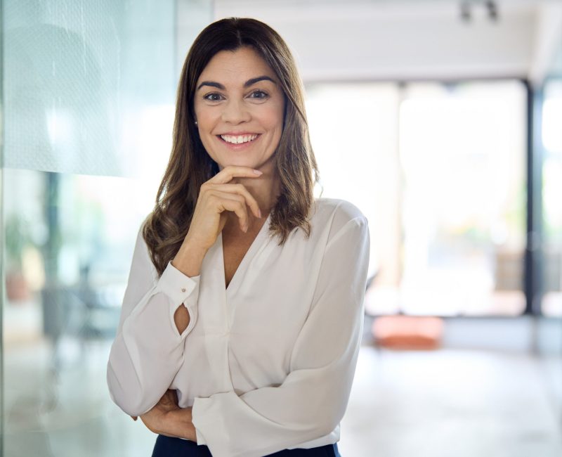 Happy confident Latin professional mid aged business woman in office, portrait. Smiling lady corporate leader, mature female executive, lady manager standing in looking at camera, portrait.