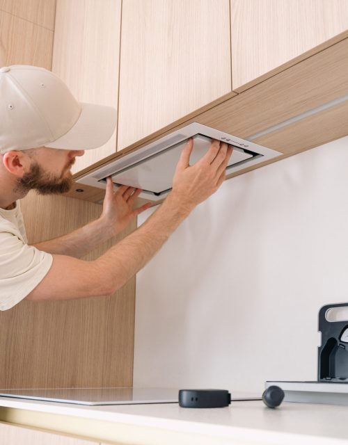 A professional handyman is focused on installing a range hood above the kitchen cabinetry in an apartment. Tools are neatly organized on the countertop, showcasing a tidy workspace.