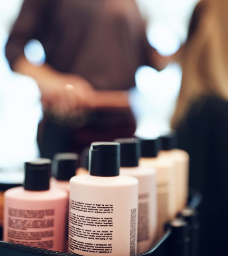 Closeup of shampoo and conditioner bottles on a tray in a hair salon with a hairdresser styling a client's hair in the background